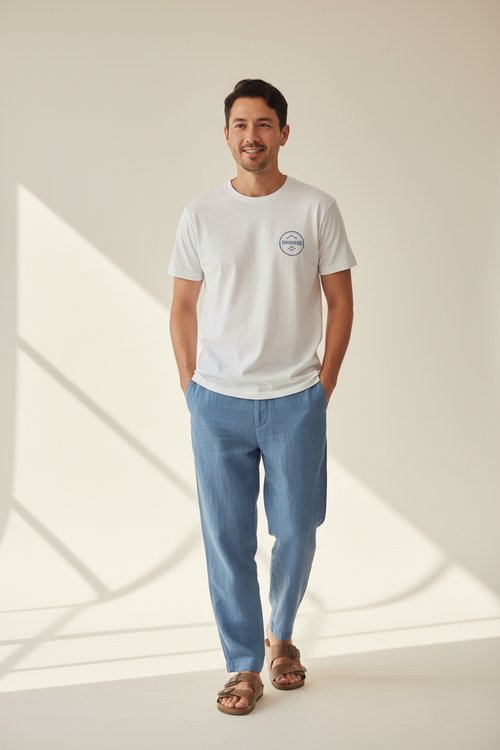 Man in White Short sleeved t-shirt with Logo "Freshwater Co." wearing blue linen pants and brown sandals. make image height 1248px and width 832px